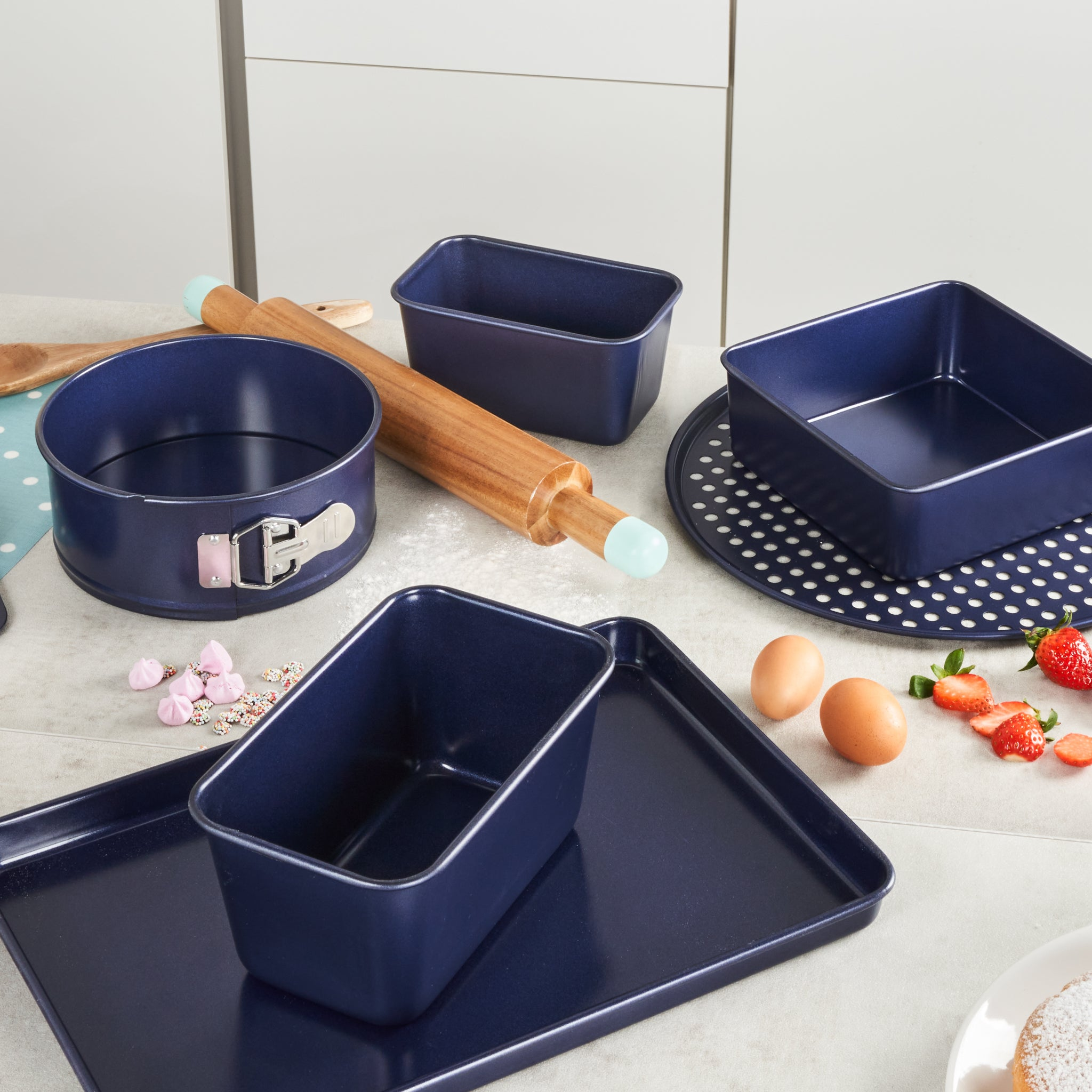 A set of blue baking pans, a rolling pin, eggs, strawberries, and scattered baking ingredients are arranged on a kitchen counter.