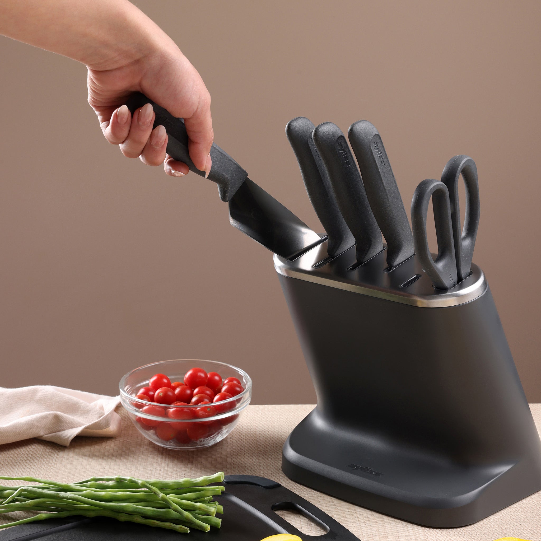 A wooden knife block holds four kitchen knives with black handles. In the background, a bowl contains lemons, fennel, and radicchio, sitting on a light countertop.
