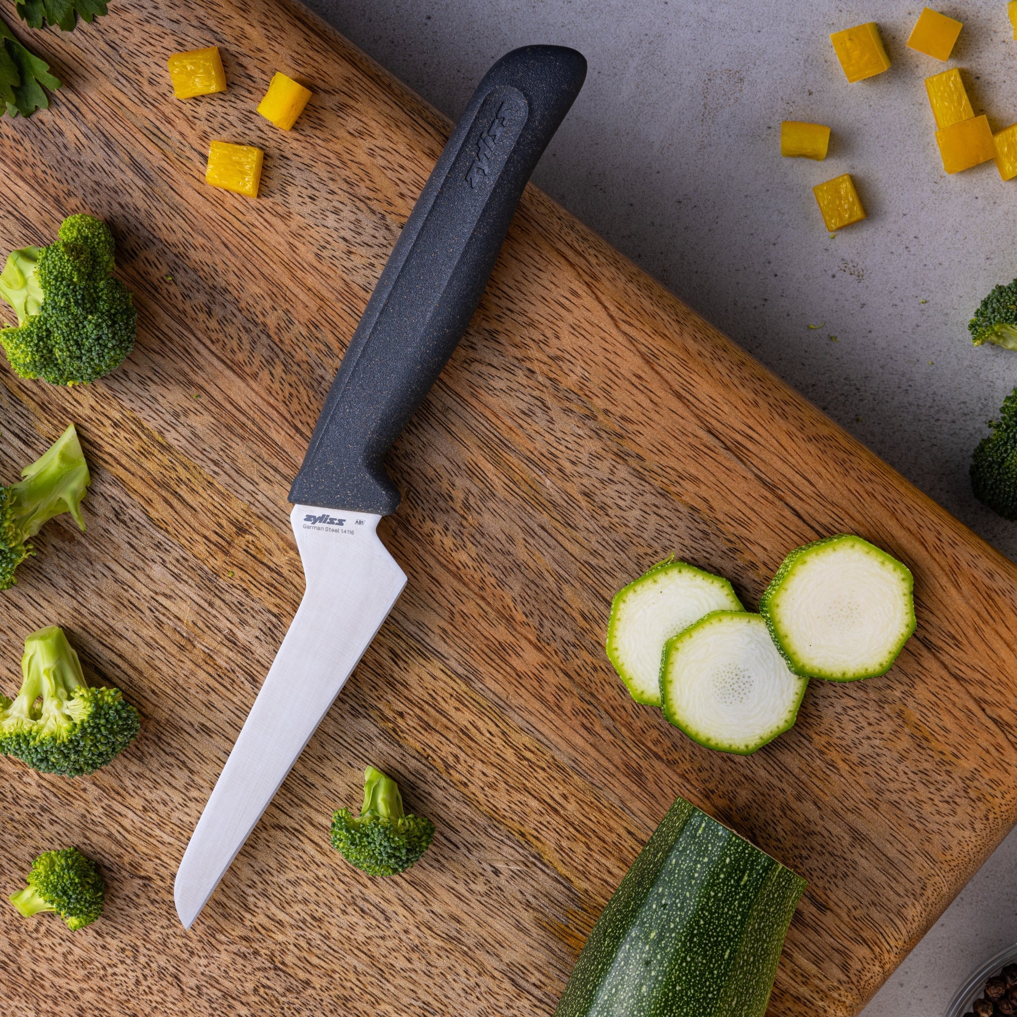 A kitchen knife on a wooden cutting board with sliced zucchini, broccoli florets, and diced yellow bell pepper scattered around.