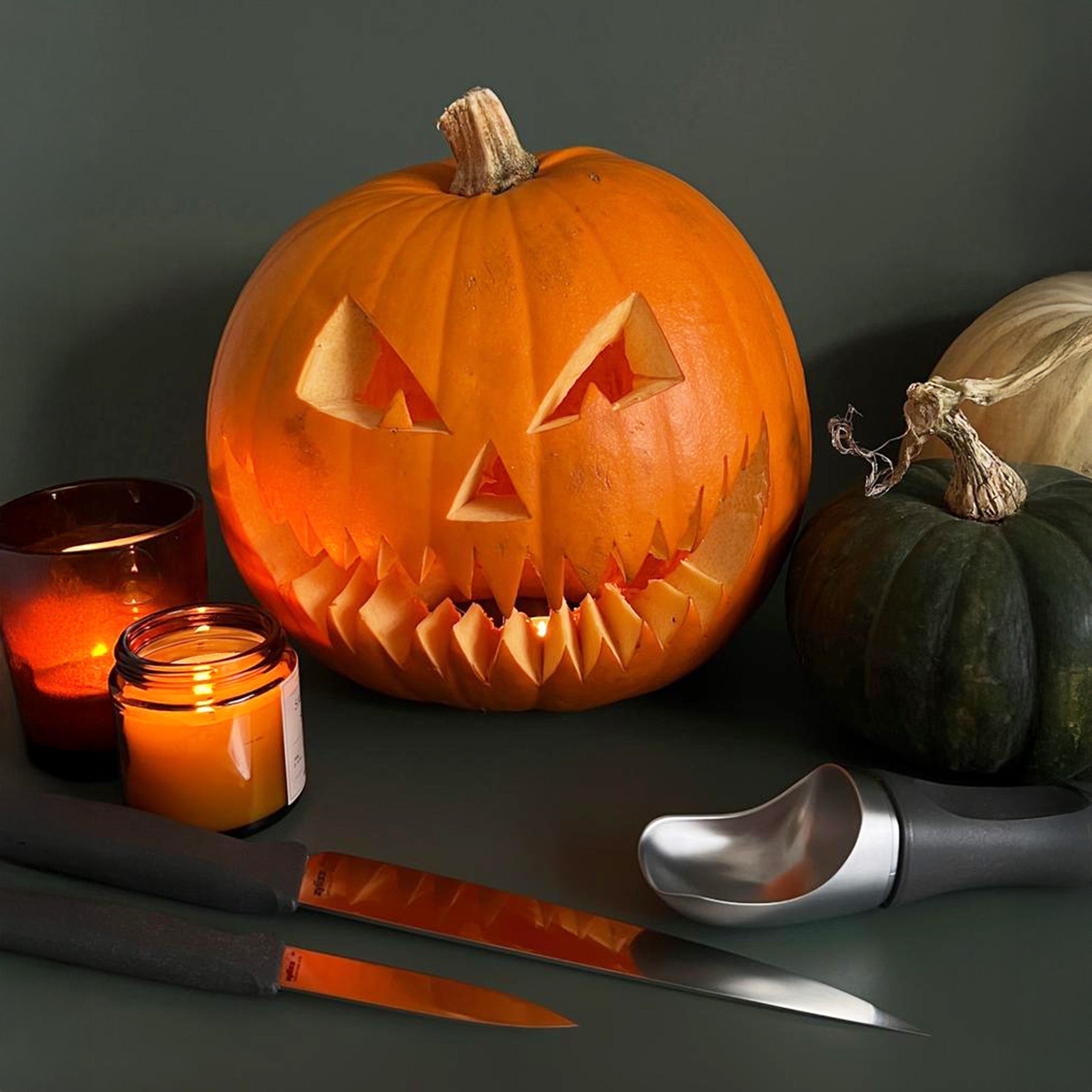 A carved jack-o’-lantern with a menacing grin is surrounded by candles, pumpkins, knives, and a carving tool, creating a spooky Halloween scene on a dark background.