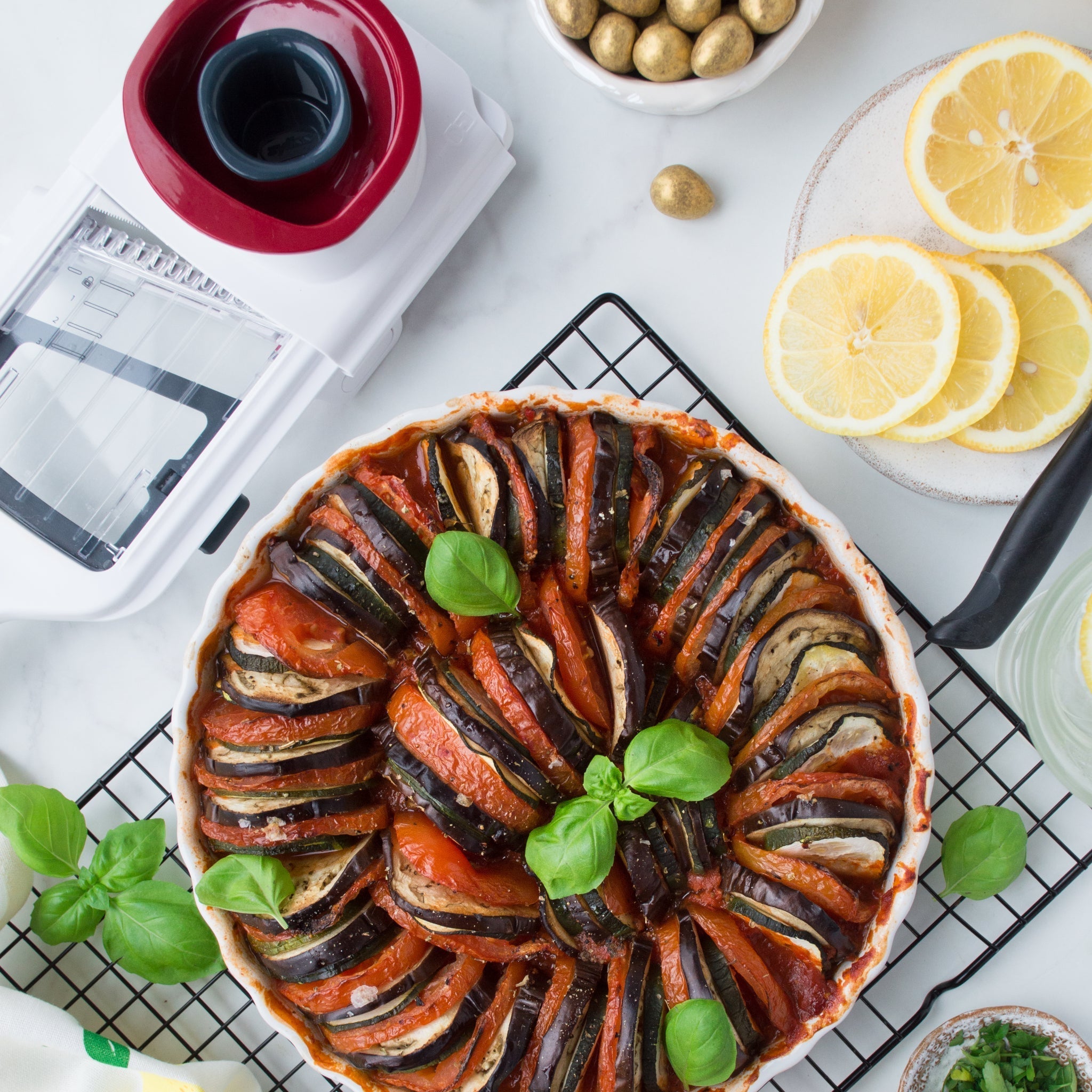 A baked ratatouille dish garnished with fresh basil sits on a cooling rack, surrounded by lemon slices, olives, and a mandoline slicer on a white countertop.