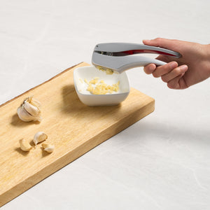 A hand uses the Zyliss Susi 3 Garlic Press with an ergonomic handle to crush garlic cloves into a small white bowl on a wooden cutting board, surrounded by whole garlic bulbs and peeled cloves. 