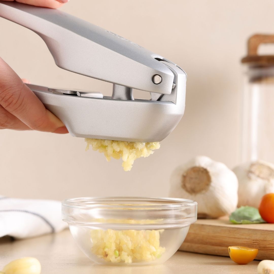 A hand uses the Zyliss Susi 4 Garlic Press with ergonomic handles to mince garlic into a glass bowl on a kitchen counter, with garlic bulbs and a yellow tomato in the background for easy cleanup.