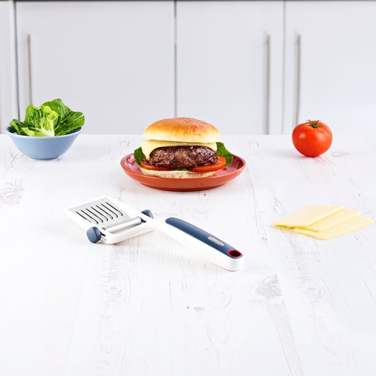 A cheeseburger sits on a white kitchen counter with a bowl of lettuce, tomato, cheese slices, and a Zyliss Dial & Slice Cheese Slicer nearby. White cabinets are in the background. 