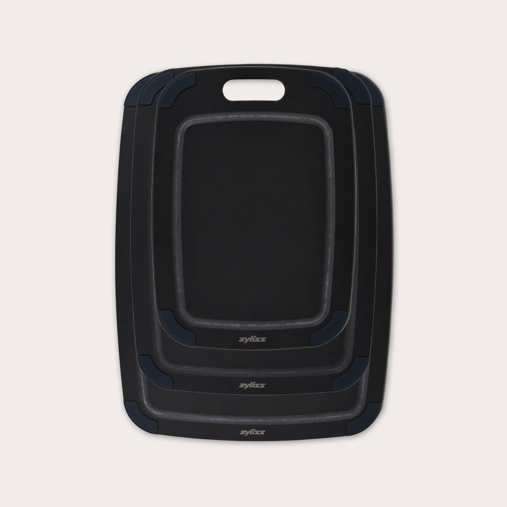 The Zyliss Wood Fibre Cutting Boards set features three nested black boards in different sizes, each with a handle slot and dark gray corner grips, made from eco-friendly recycled wood fibres and shown on a light background. #set of 3