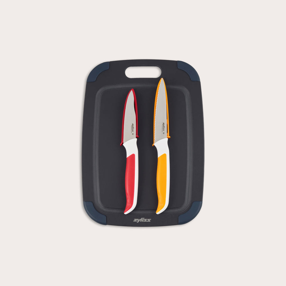 The Zyliss 3 Piece Wood Fibre Chopping Board & Knife Set includes a black cutting board with rounded corners and two knives—one red-handled and one orange-handled—both with silver blades. 