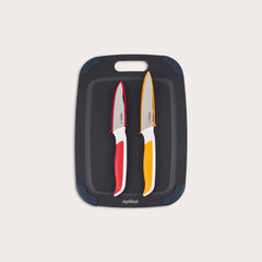 The Zyliss 3 Piece Wood Fibre Chopping Board & Knife Set includes a black cutting board with rounded corners and two knives—one red-handled and one orange-handled—both with silver blades. 