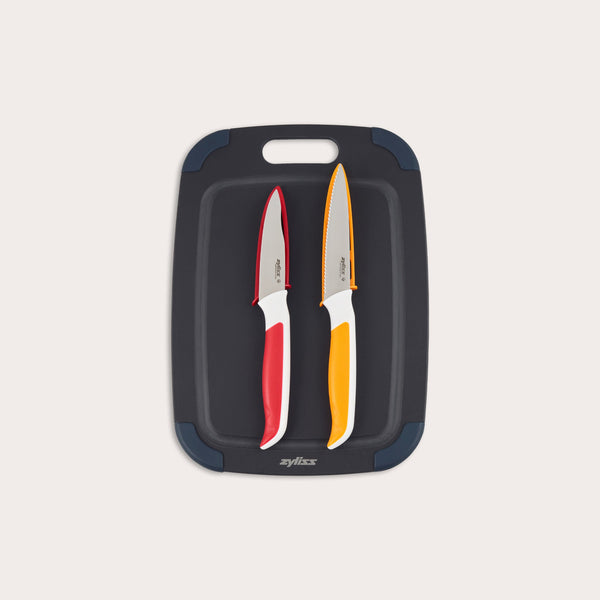 The Zyliss 3 Piece Wood Fibre Chopping Board & Knife Set includes a black cutting board with rounded corners and two knives—one red-handled and one orange-handled—both with silver blades. 