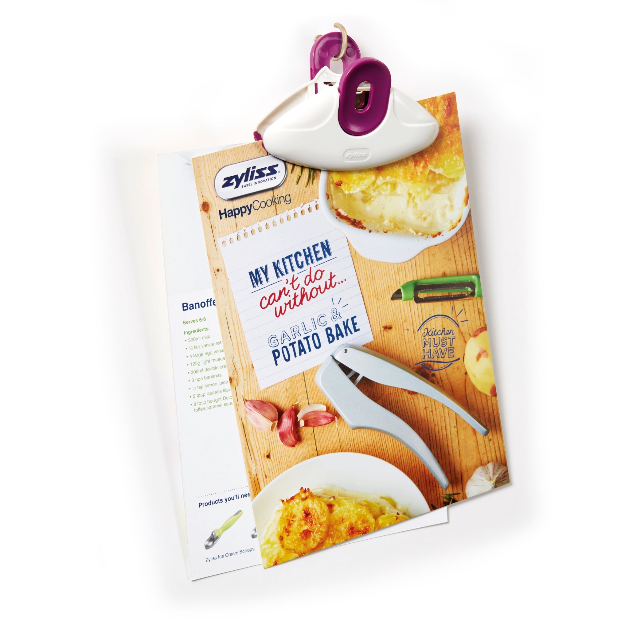 A Zyliss Clip-All Bag Clip (purple and white), a kitchen must-have, secures a garlic & potato bake recipe page with photos of baked dishes, garlic cloves, a peeler, and the line: My kitchen cant do without... GARLIC & POTATO BAKE. 