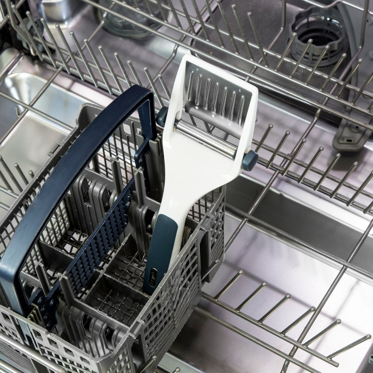 A vegetable peeler with a blue and white handle is placed inside the cutlery basket of an open, empty dishwasher.