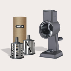 #drumgraterset The Zyliss Gourmet Drum Grater, made of gray stainless steel with a hand crank and two interchangeable blades, comes in a brown cardboard tube labeled Zyliss and is shown on a white background.