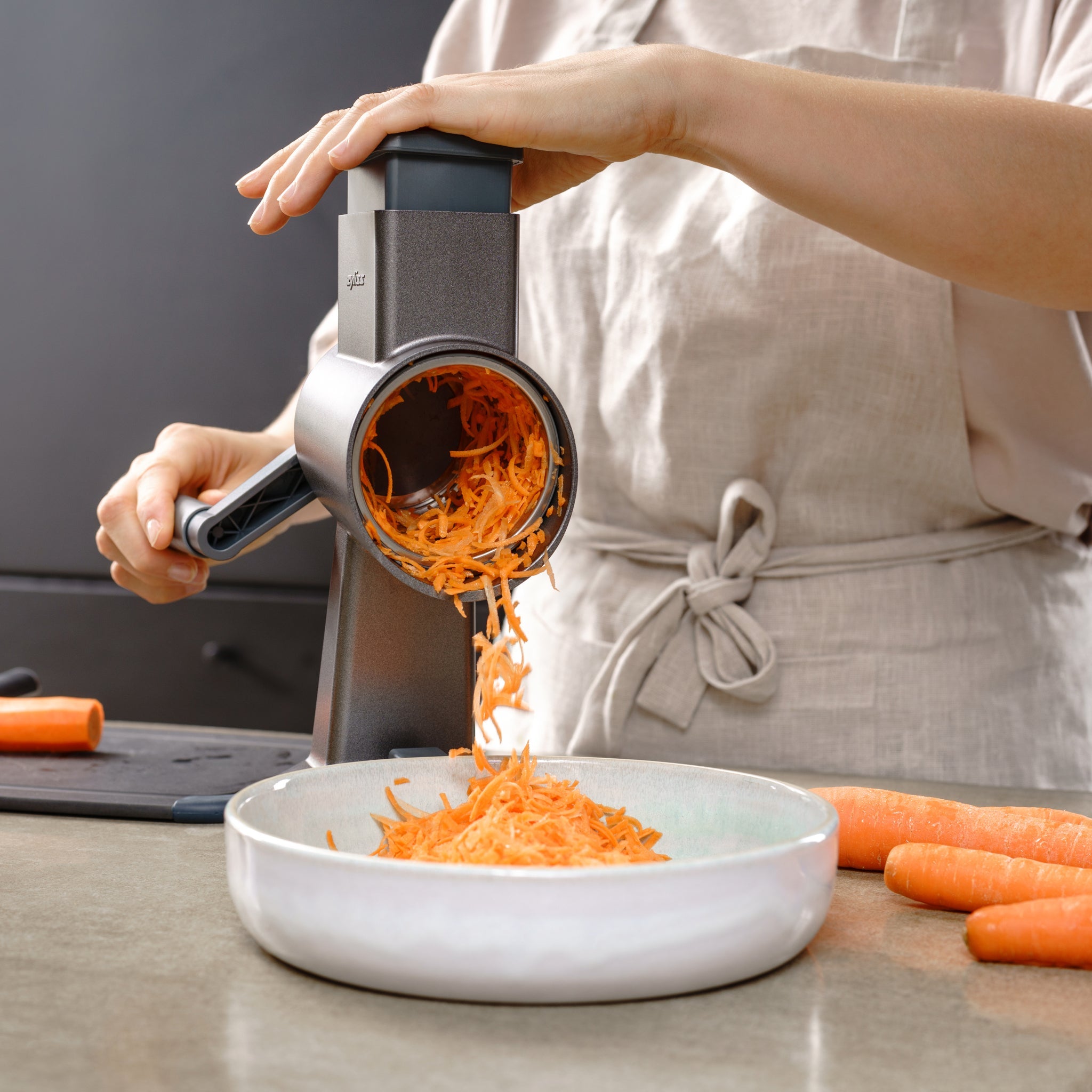 Wearing a beige apron, someone uses the Zyliss Julienne Drum Gourmet to shred carrots into a white bowl, with whole carrots and a cutting board on the counter. 