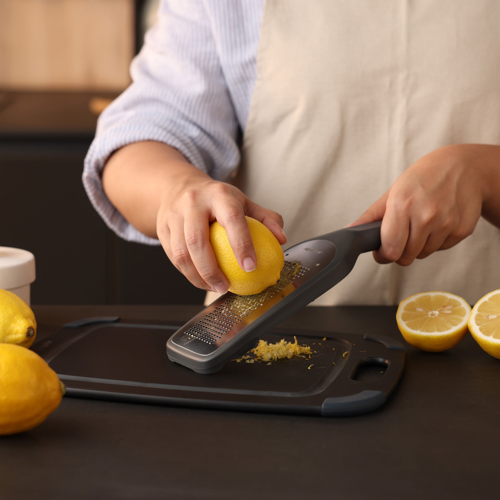 A person wearing an apron is zesting a lemon using a grater over a cutting board, with whole and halved lemons nearby on a kitchen counter.