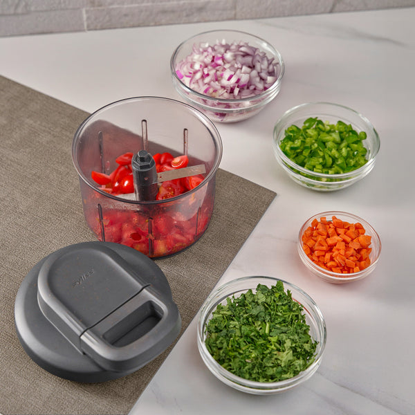 The Zyliss Easy Pull Food Processor, filled with chopped tomatoes, sits on a table with bowls of red onion, green bell pepper, carrot, and cilantro. Its pull cord and lid are placed nearby for easy use.