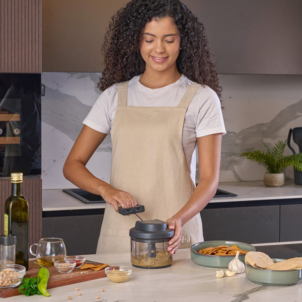 A woman in a beige apron uses the Zyliss Easy Pull Food Processor in a modern kitchen, with chickpeas, bread, and garlic on the countertop beside her.