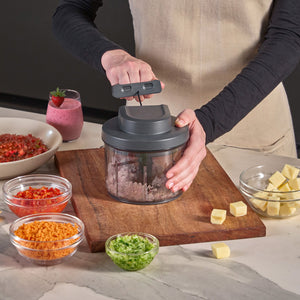 Someone uses the Zyliss Easy Pull Food Processor on a wooden cutting board, surrounded by bowls filled with chopped veggies, cheese cubes, and a pink smoothie on the kitchen counter.
