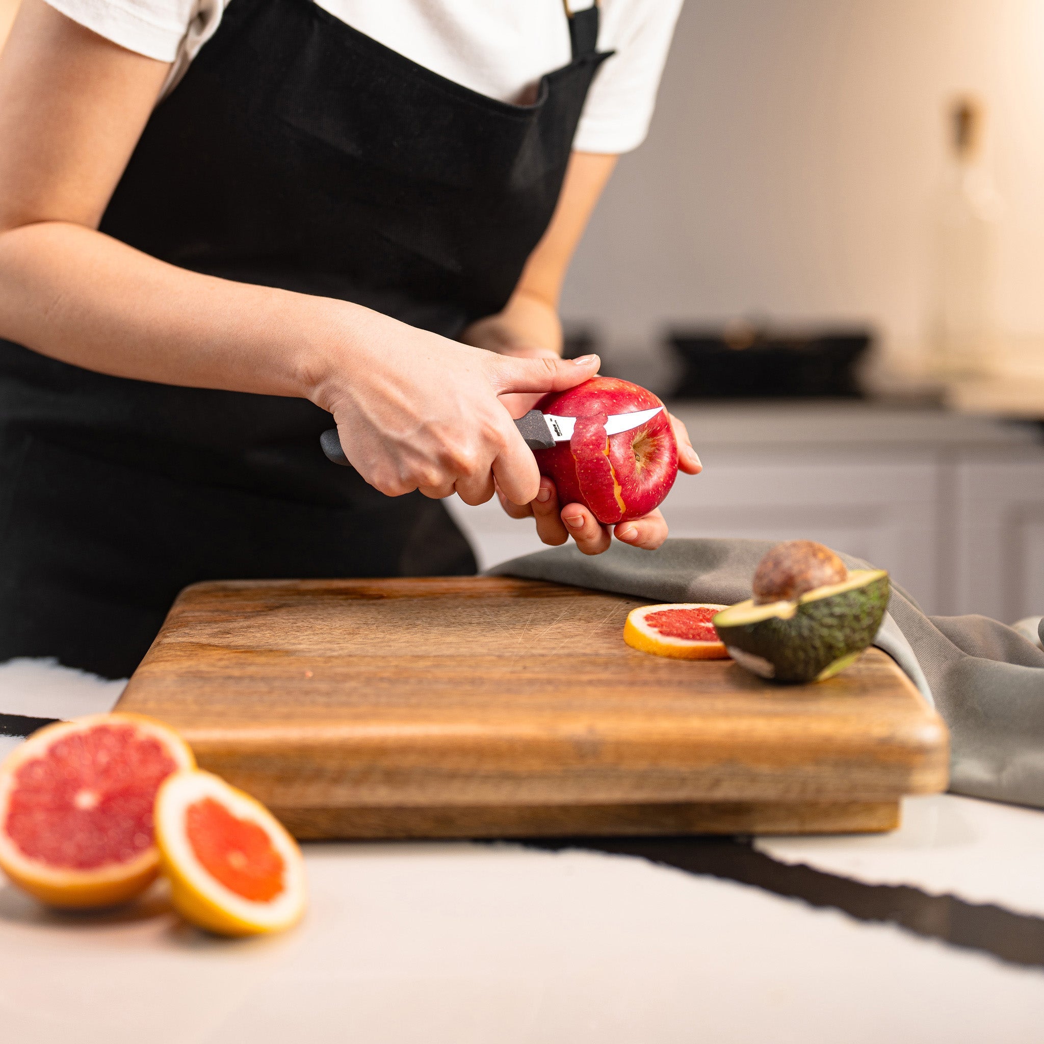 Wearing a black apron, someone peels a pomegranate with the Zyliss Peeling Knife 6cm / 2½ over a wooden cutting board in the kitchen, next to sliced grapefruit and avocado. 