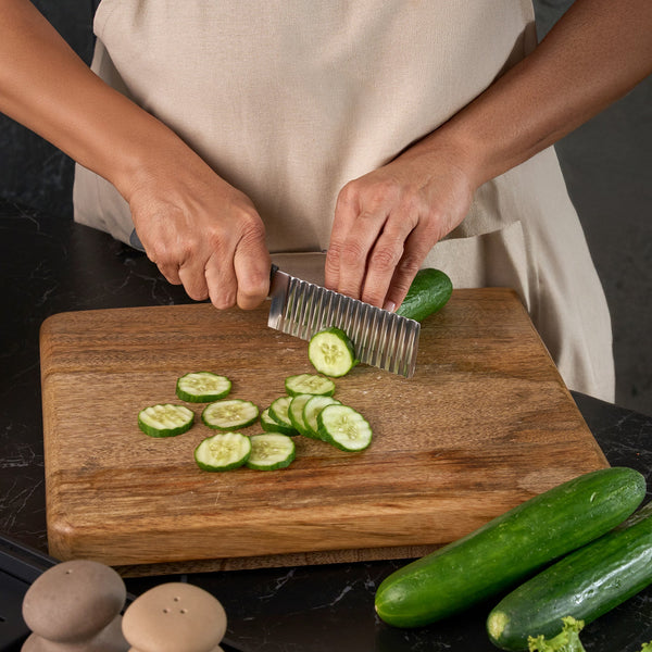 Wearing a beige apron, a person slices a cucumber with the Zyliss Crinkle Cut Knife 11.5cm/4" on a wooden board. Cucumber slices, whole cucumbers, and salt and pepper shakers are also present on the board.