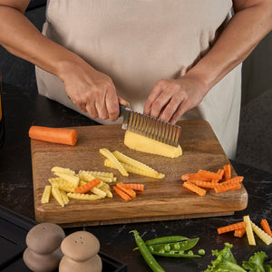 A person slices potatoes with the Zyliss Crinkle Cut Knife 11.5cm/4" on a wooden board. Crinkle-cut potato and carrot pieces, a whole carrot, green beans, and lettuce are also on the board.