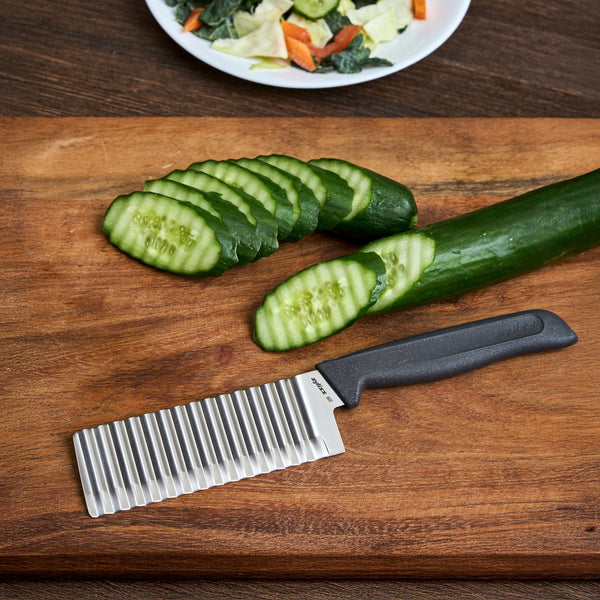 The Zyliss Crinkle Cut Knife 11.5cm/4" with an ergonomic handle and stainless steel blade lies next to a crinkle-cut cucumber on a wooden board, with a plate of mixed vegetables in the background.
