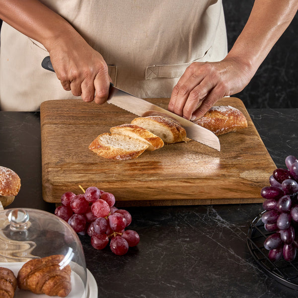 Wearing a beige apron, a person slices a baguette on a wooden board using the Zyliss Bread Knife 21cm/8". Red grapes are nearby, croissants sit under a glass dome to the left, and more grapes rest on a black wire rack to the right.