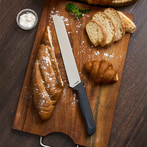 A sliced baguette and a croissant sit on a wooden board with the Zyliss Bread Knife 21cm/8", featuring a serrated stainless steel blade. Nearby are a bowl of cream and a basket of sliced bread, all placed on a dark wooden surface.