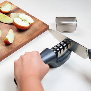 A person uses the Zyliss Knife Sharpener on a white surface to maintain kitchen blades, with sliced apples on a wooden cutting board nearby.