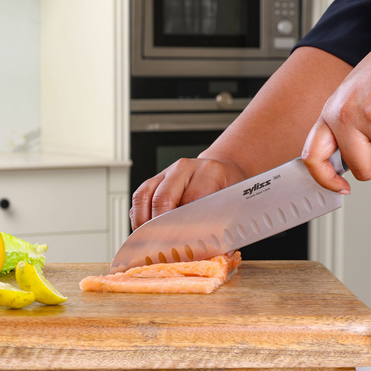 A person slices salmon on a wooden cutting board using the Zyliss Comfort Pro Santoku Knife 18.5cm/7" with an ergonomic handle. Slices of lemon and lettuce are nearby, set in a modern kitchen backdrop.