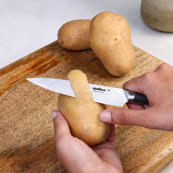 A person peels a potato with the Zyliss Comfort Pro Utility Knife 14cm / 5½" in one hand, while holding the potato in the other. Two unpeeled potatoes and a container rest on a wooden cutting board in the background.