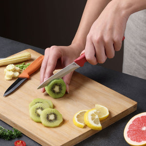 A person slices a kiwi on a wooden cutting board using the Zyliss Classic 2 Piece Paring Knife Set with an ergonomic handle. Slices of lemon, baby corn, and half a grapefruit are also visible on the board and table.