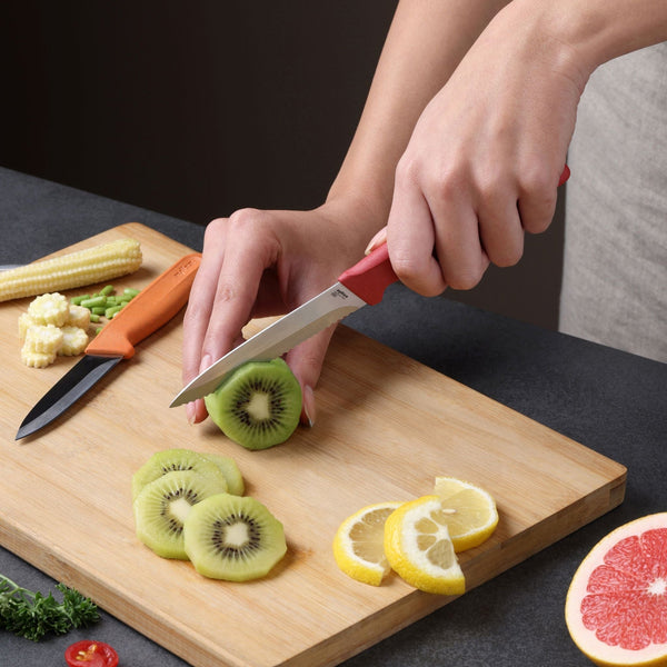 A person slices a kiwi on a wooden cutting board using the Zyliss Classic 2 Piece Paring Knife Set with an ergonomic handle. Slices of lemon, baby corn, and half a grapefruit are also visible on the board and table.