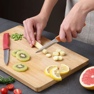 A person slices baby corn on a wooden cutting board with a Zyliss Classic 2 Piece Paring Knife Set featuring ergonomic handles and sharp stainless steel blades. Sliced kiwi, lemon, green pepper, cherry tomatoes, and grapefruit are nearby.