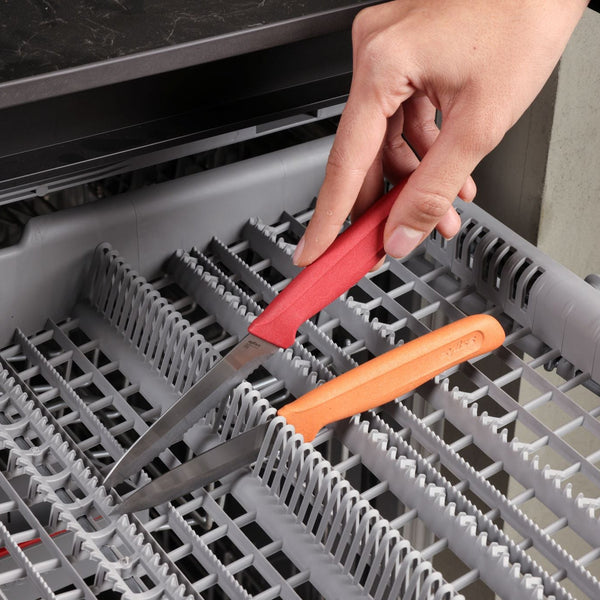 A hand places two Zyliss Classic Paring Knives with ergonomic handles and stainless steel blades blade-down into the cutlery rack of an open dishwasher.