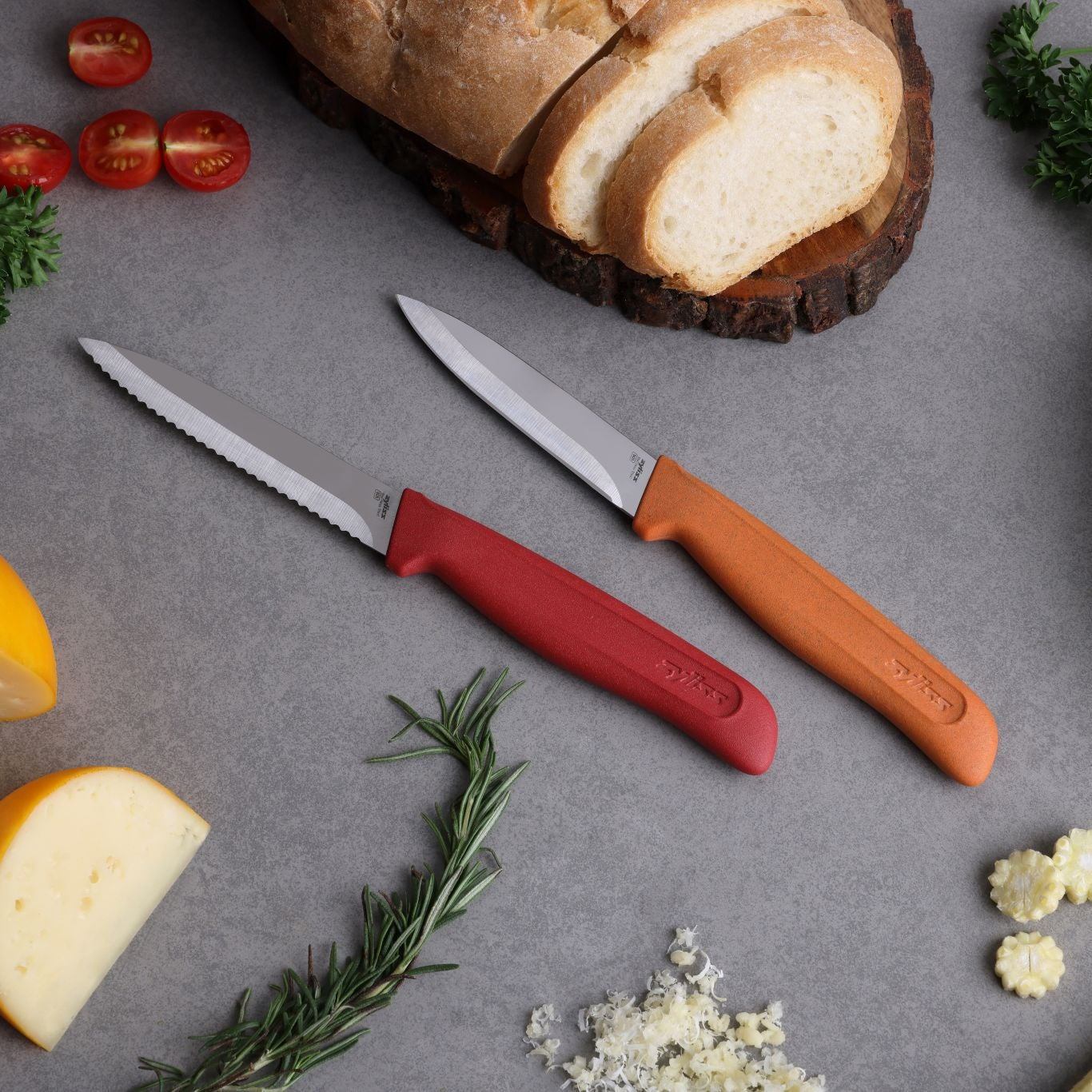 The Zyliss Classic 2 Piece Paring Knife Set, featuring ergonomic handles and stainless steel blades, rests on a gray countertop with sliced bread, cheese, herbs, cherry tomatoes, and minced garlic nearby.