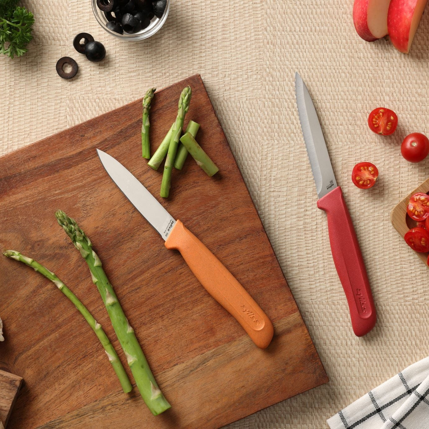 The Zyliss Classic 2 Piece Paring Knife Set features ergonomic handles and stainless steel blades, displayed on a wooden surface with asparagus, tomatoes, olives, apple slices, and a checkered napkin on a beige textured background.