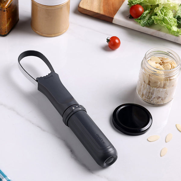 A Zyliss Jar Opener with an adjustable rubberized band and looped handle sits on a white countertop near a jar of sliced almonds, tomato, lettuce, and a black lid. Kitchen items and food are visible in the background. 