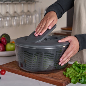 On a kitchen counter, a person uses the Zyliss Swift Dry Salad Spinner with its one-handed pump surrounded by fresh veggies like lettuce, tomatoes, and avocados, while wine glasses hang in the background.