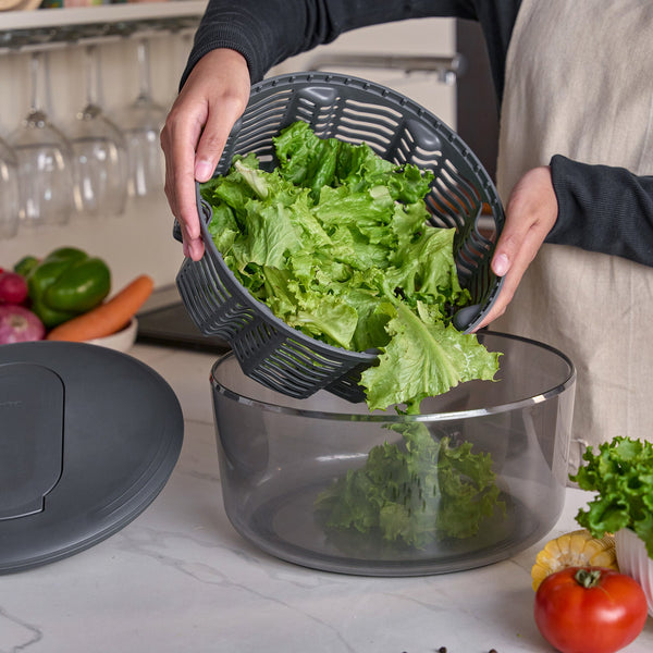 Using the Zyliss Swift Dry Salad Spinner’s one-handed pump, a person dries lettuce, then transfers crisp leaves into a salad bowl on the kitchen counter amid fresh vegetables and the spinner lid.