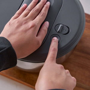 A person presses the button on the Zyliss Swift Dry Salad Spinner Stainless Steel lid while steadying it with their other hand, similar to using a one-handed pump; the appliance rests on a wooden surface.