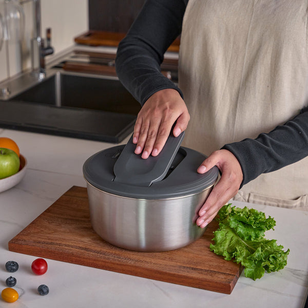 A person in a beige apron places the gray lid on the Zyliss Swift Dry Salad Spinner Stainless Steel next to a stainless steel bowl on a wooden cutting board, with lettuce and assorted fruits nearby on the kitchen counter.