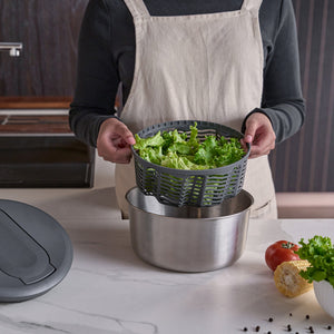 In a kitchen, someone in an apron holds a basket of lettuce above the Zyliss Swift Dry Salad Spinner Stainless Steel, with fresh vegetables like tomato and corn visible on the counter.
