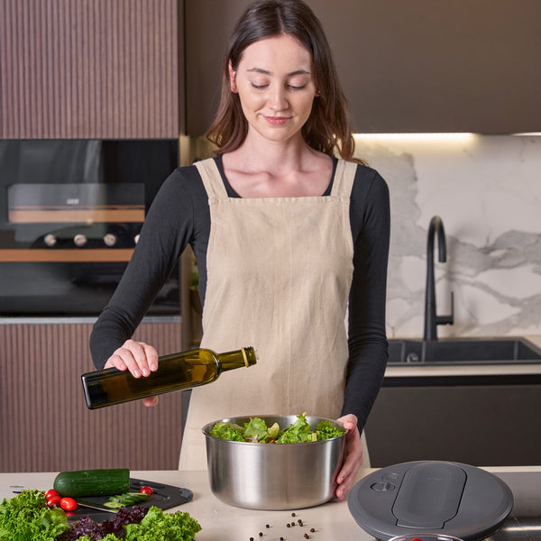 In a modern kitchen, a woman in a beige apron adds olive oil to fresh salad. Nearby, the Zyliss Swift Dry Salad Spinner Stainless Steel sits on the counter with assorted vegetables, a stainless steel bowl, and other kitchen appliances.