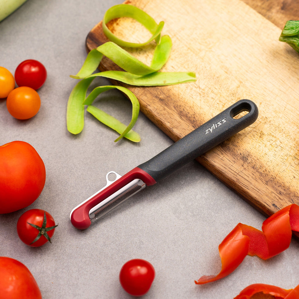 The Zyliss Soft Skin Peeler features a dark gray handle with a small end hole, a red top, and a horizontal stainless steel blade. This eco-friendly peeler is displayed on a plain light background. 