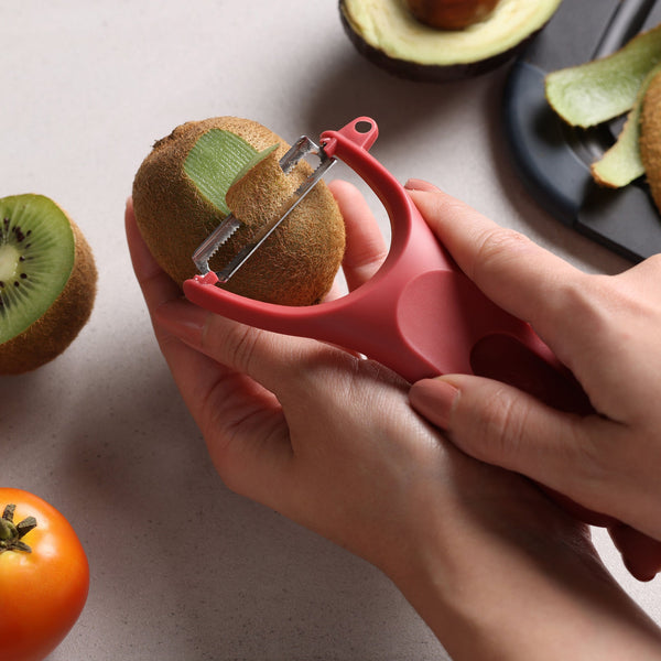 A person uses a red peeler from the Zyliss 3 Piece Y Peeler Set to peel a kiwi, with sliced avocado, tomato, and a black cutting board in the background.