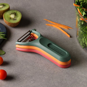 The Zyliss 3 Piece Y Peeler Set in green, orange, and pink rests on a gray countertop beside sliced kiwi, carrot sticks, leafy greens, cherry tomatoes, and cucumber slices.