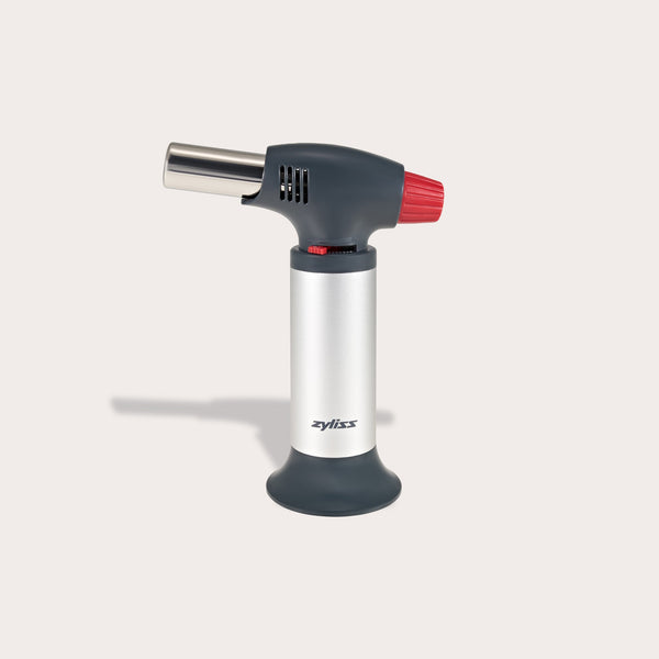 The Zyliss Kitchen Blowtorch features a metallic body, ergonomic gray handle, and red ignition switch. It stands upright on a light surface and offers adjustable flame control for precise kitchen use. The brand name is visible on the front. 