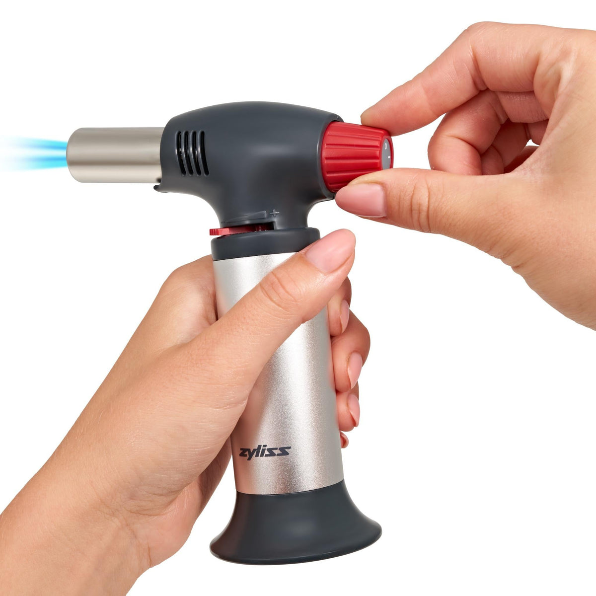 A person holds a silver and black kitchen torch, turning the red ignition knob with one hand while a blue flame comes out of the nozzle. The torch is branded Zyliss.