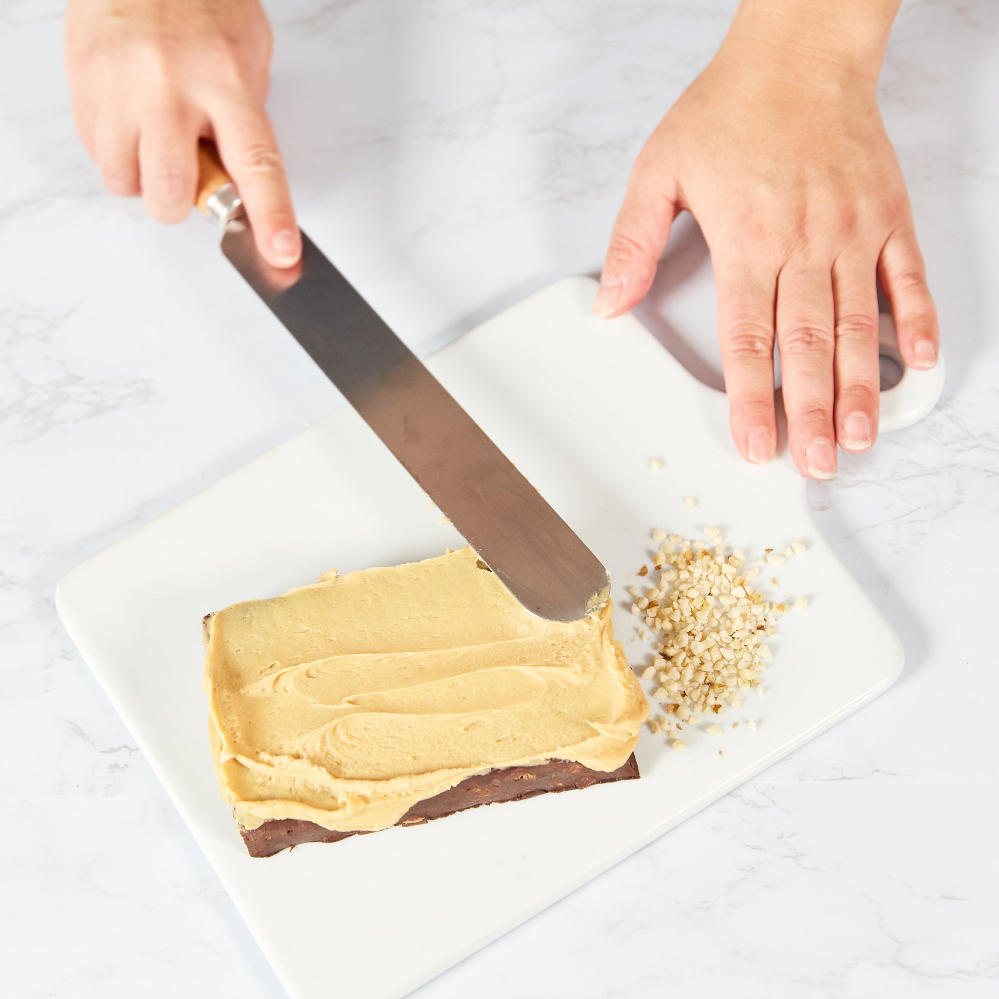 Someone spreads creamy frosting on a rectangular brownie using the Zyliss Beech Wood Palette Knife beside a small pile of finely chopped nuts on a white cutting board. 