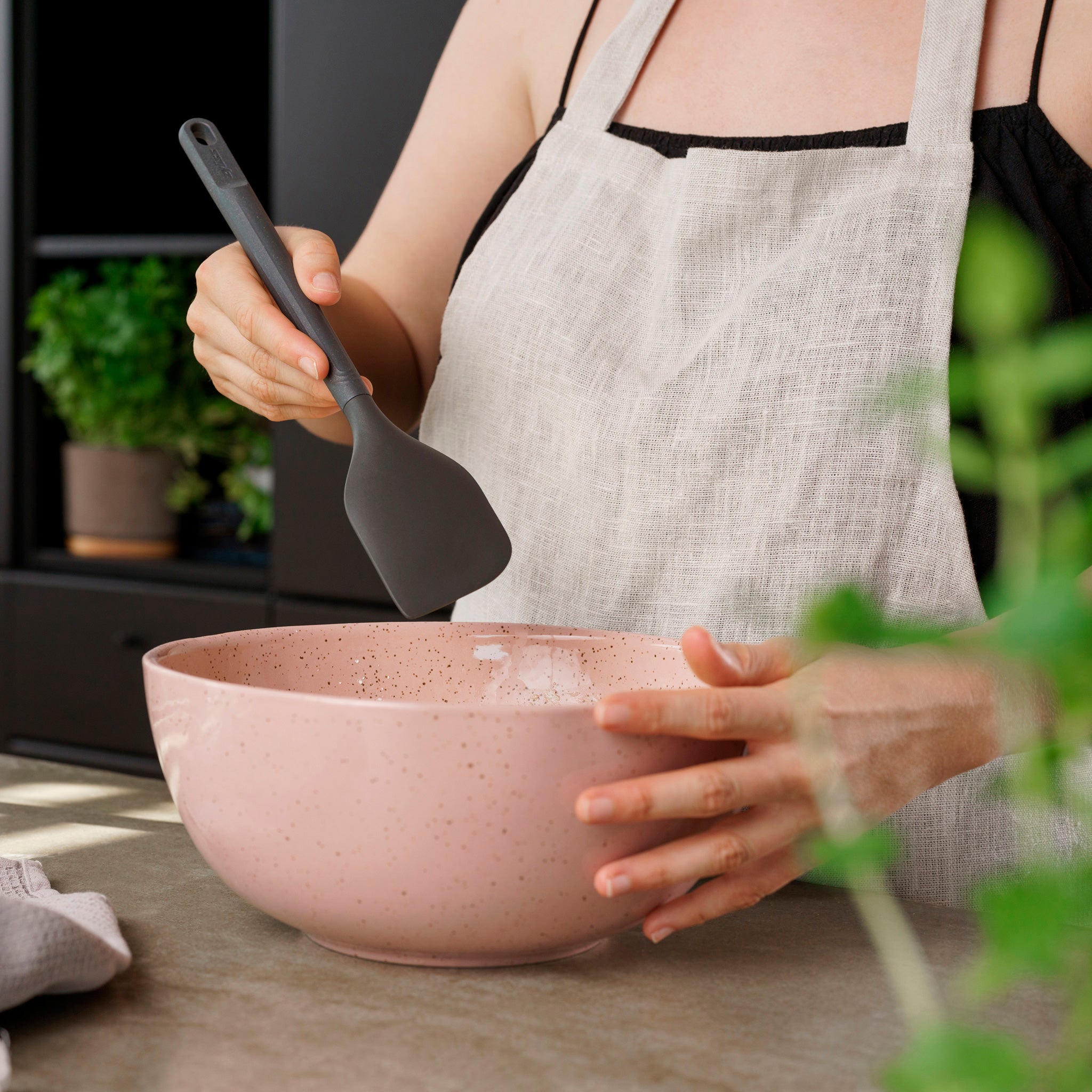 Wearing a light apron, a person stands at a kitchen counter with green plants in the background, stirring a pink bowl with a Zyliss Spatula. #Large