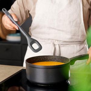 A person in a light-colored apron holds a Zyliss Square Mixing Spoon above a saucepan of orange soup on the stove, with only their torso and hands visible. 